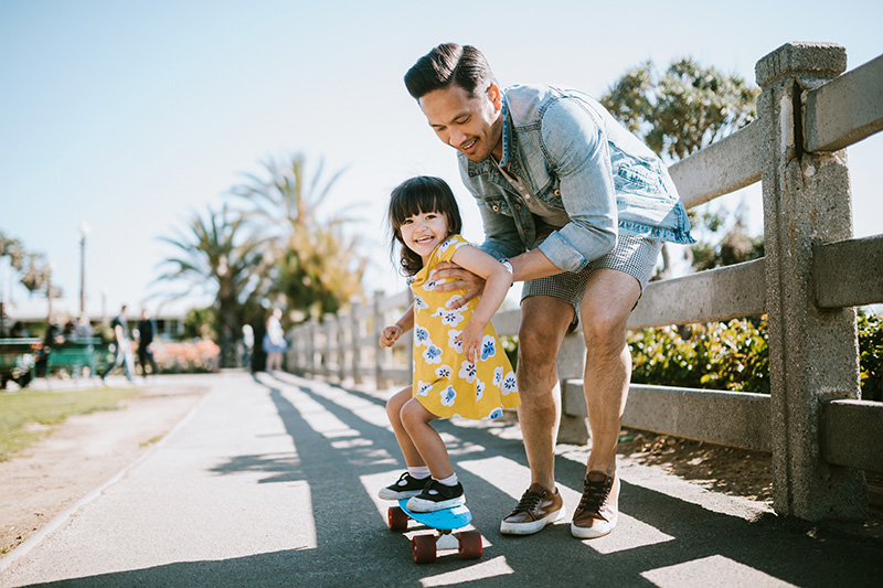 Un papa aide sa petite fille à faire du skateboard, en la soutenant par la taille. Filmé à Los Angeles, Californie, près de la jetée de Santa Monica.