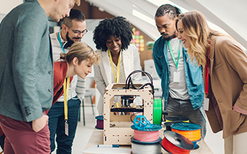 L'image montre un groupe de personnes rassemblées autour d'une imprimante 3D, observant son fonctionnement.