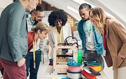 The image shows a group of people gathered around a 3D printer, observing its operation.