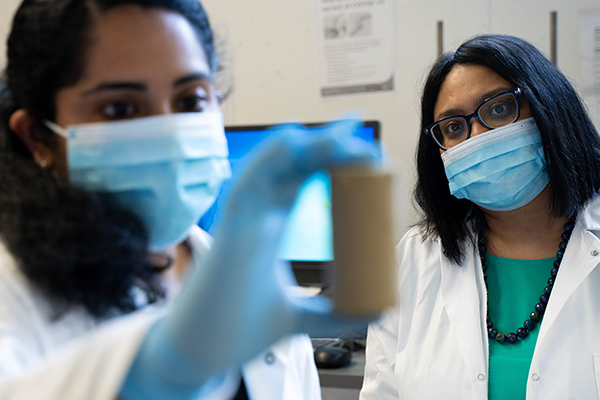 Two scientists wearing masks and lab coats examine a sample in a laboratory.