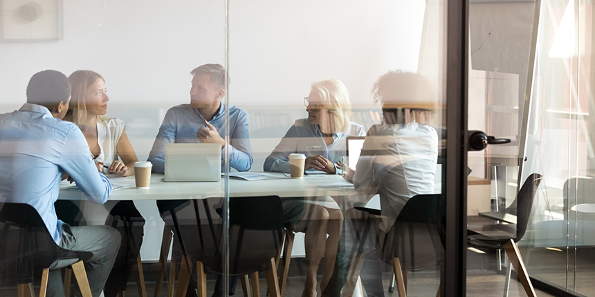 Five people are sitting around a table in a glass-walled office room, engaged in a meeting with documents, laptops, and coffee cups in front of them.