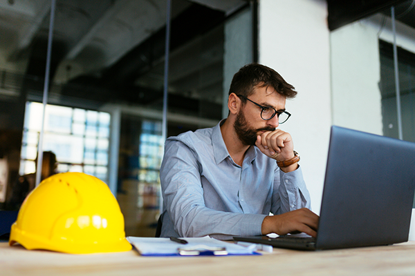 A man with glasses sits at a desk, focused on his laptop. A yellow hard hat, papers, and a pen are on the desk.