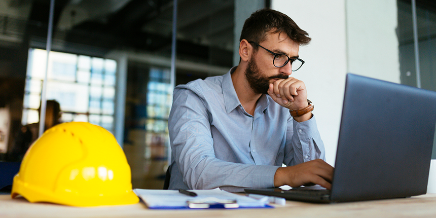 A man with glasses sits at a desk, focused on his laptop. A yellow hard hat, papers, and a pen are on the desk.