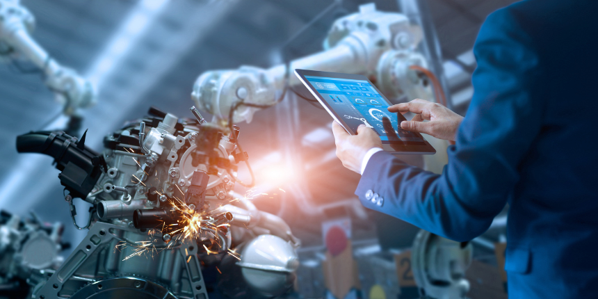 Une personne en costume utilise une tablette tactile pour contrôler des bras robotisés travaillant avec des machines, des étincelles étant visibles pendant le processus de fabrication.