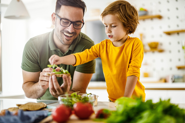 Père et fils préparant joyeusement une salade dans une cuisine lumineuse. Le père en lunettes tient un bol, tandis que le fils en chemise jaune ajoute des feuilles, entouré de légumes frais.