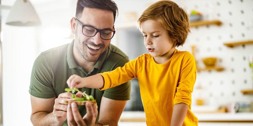 Father and son joyfully preparing a salad in a bright kitchen. The father in glasses holds a bowl, while the son in a yellow shirt adds leaves, surrounded by fresh veggies.