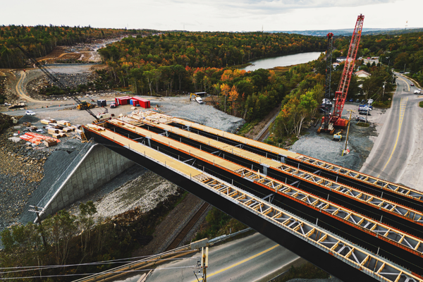 Aerial view of a bridge under construction spanning across a road, with cranes, construction materials, and a river surrounded by trees in the background.