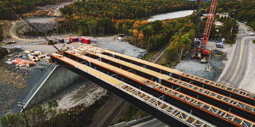 Aerial view of a bridge under construction spanning across a road, with cranes, construction materials, and a river surrounded by trees in the background.