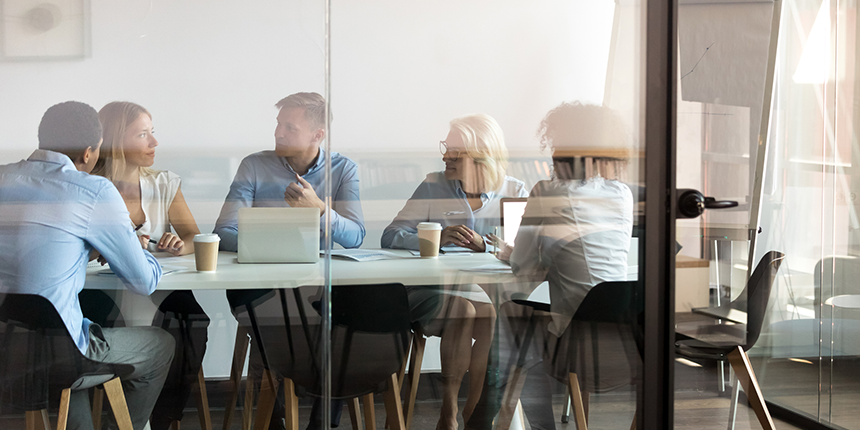 Cinq personnes sont assises autour d'une table de conférence dans un bureau aux murs vitrés, engagées dans une discussion, avec leurs ordinateurs portables, des documents et des tasses de café devant elles.
