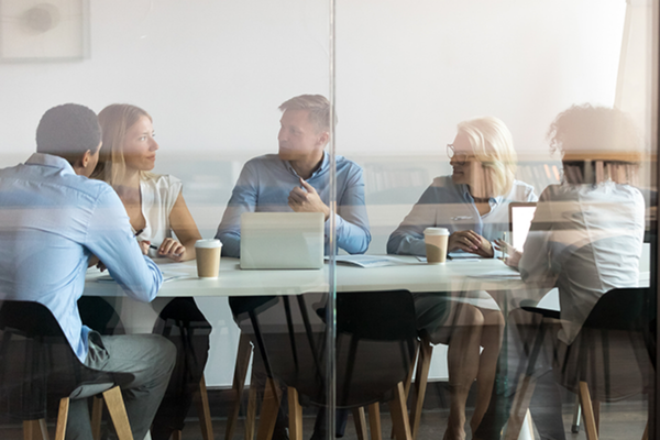 Five people sit around a conference table in a glass-walled office, engaged in discussion with laptops, papers, and coffee cups in front of them.