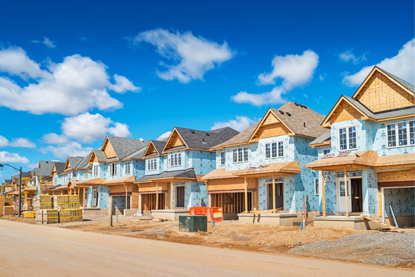 A row of partially constructed suburban houses with blue siding under a clear blue sky. Construction materials and equipment are visible in the foreground.