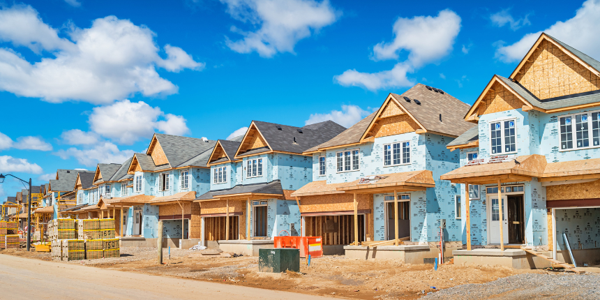 A row of partially constructed suburban houses with blue siding under a clear blue sky. Construction materials and equipment are visible in the foreground.