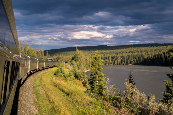A scenic train winds along a lush green forest beside a calm river under a dramatic cloudy sky, evoking a sense of adventure and tranquility.