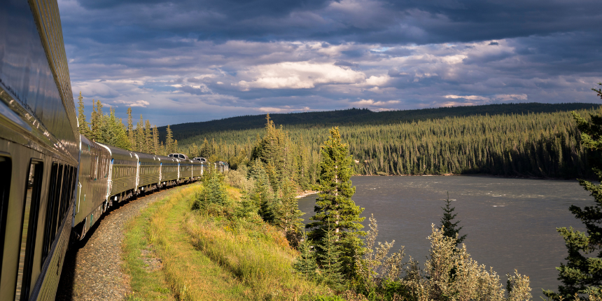 A scenic train winds along a lush green forest beside a calm river under a dramatic cloudy sky, evoking a sense of adventure and tranquility.