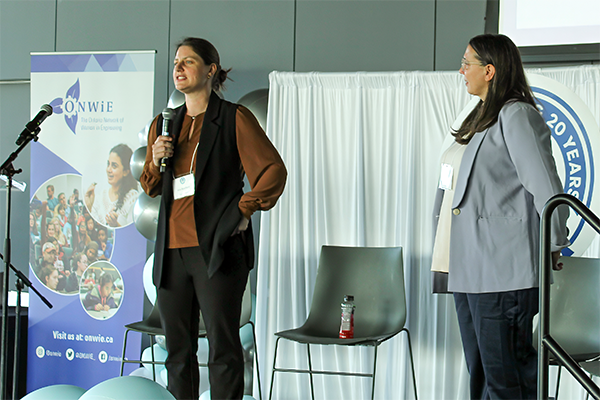 Two women stand on a stage speaking at a conference; one holds a microphone while the other listens. A banner for ONWiE is visible in the background.