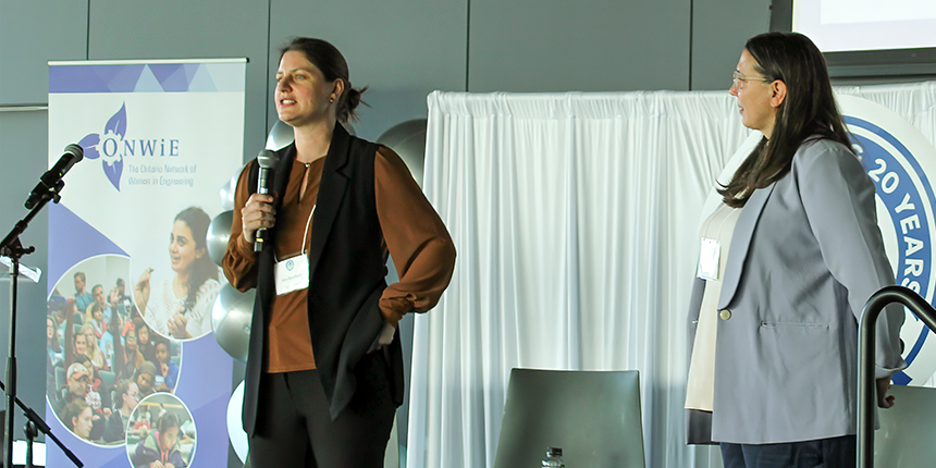 Two women stand on a stage speaking at a conference; one holds a microphone while the other listens. A banner for ONWiE is visible in the background.