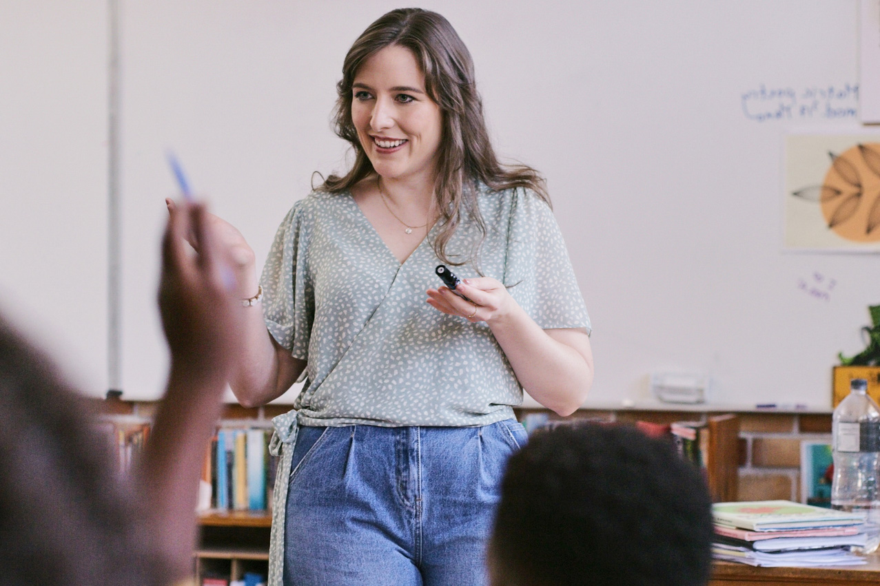 Teacher standing at the front of a classroom with a student raising their hand to ask a question