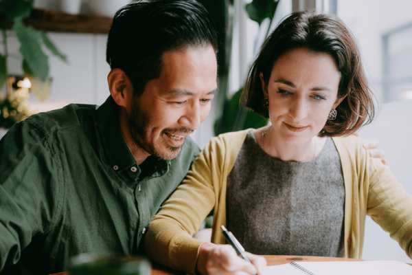  Gemini said A man and a woman sit closely together at a table, smiling as they focus on writing in a notebook