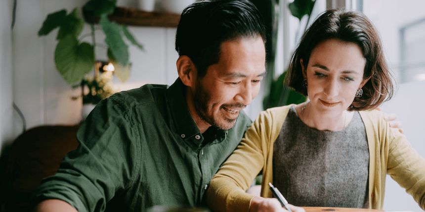 A man and a woman sit closely together at a table, smiling as they focus on writing in a notebook.