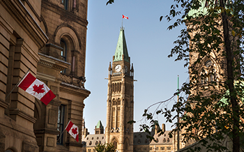 Une vue de la Tour de la Paix sur la Colline du Parlement à Ottawa, encadrée par des bâtiments historiques en pierre et des drapeaux canadiens sous un ciel bleu clair.