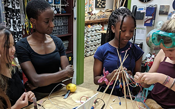 Une éducatrice observe trois jeunes élèves travaillant ensemble sur un projet scientifique pratique impliquant des tiges en bois, des fils et un citron dans une salle de classe ou un atelier.