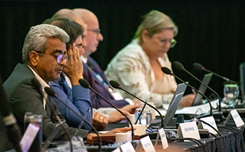 Un groupe de quatre professionnels assis à une longue table lors d'une conférence. Ils utilisent des microphones et des ordinateurs portables tout en écoutant une présentation ou une discussion.