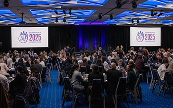 Une grande salle de conférence remplie de participants assis à des tables rondes. Deux grands écrans à l'avant affichent le logo « Conférence 30 d'ici 2030 2025 » sous un plafond éclairé en bleu.