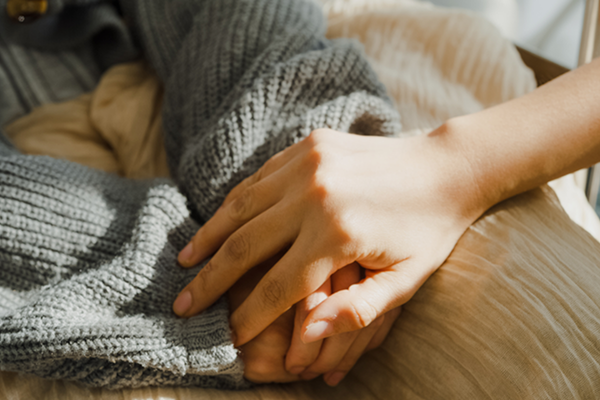 A close-up shot of one person's hand gently resting on top of another's in a gesture of comfort and support. The person being comforted is wearing a thick, grey knitted sweater.