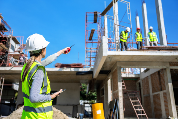 A female construction supervisor in a white hard hat and yellow safety vest points toward a building under construction while holding a radio. Three other workers in safety gear are visible on an upper level of the concrete and brick structure under a clear blue sky.