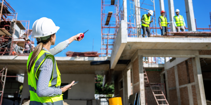 A female construction supervisor in a white hard hat and yellow safety vest points toward a building under construction while holding a radio. Three other workers in safety gear are visible on an upper level of the concrete and brick structure under a clear blue sky.