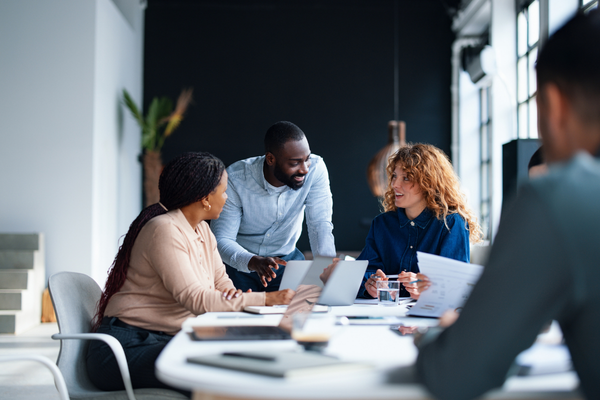 A group of diverse colleagues collaborating in a bright, modern office space. One man stands leaning over a conference table to speak with two seated women, while open laptops and printed documents are spread out in front of them.