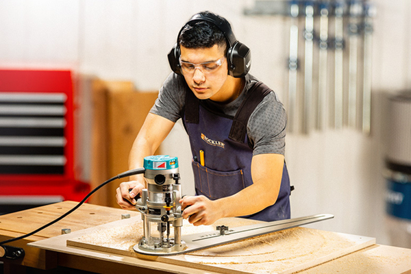 A woodworker wearing safety glasses, ear protection, and an apron operates a plunge router with a circle-cutting jig on a workbench. Sawdust covers the piece of wood as he carefully guides the tool in a workshop setting.
