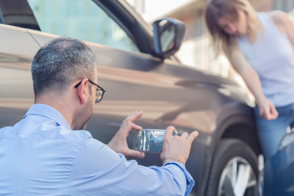 A man crouches down to take photos of vehicle damage with his smartphone after a minor car accident. A woman stands in the background looking distressed near the point of impact.