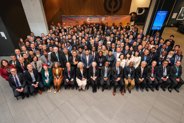 Large group photograph of delegates gathered indoors for the International Engineering Alliance event. Participants are arranged in multi-tiered rows in a modern atrium, with the front row seated. A large banner bearing the alliance logo stands in the background.