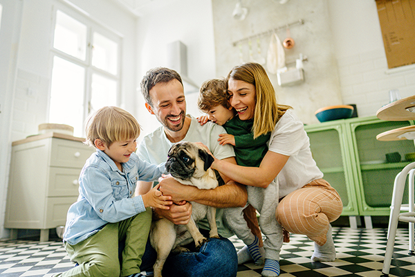 Photograph of a happy family gathered together on the tiled floor of a bright kitchen. A father, mother, and two young children surround and pet a small pug dog, smiling and laughing together.
