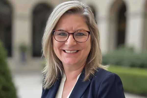 A professional portrait of  Darlene Spracklin-Reid, Engineers Canada President, smiling outdoors. She wears glasses with tortoiseshell frames, a dark blazer, and a white top, set against a softly blurred courtyard and building facade.