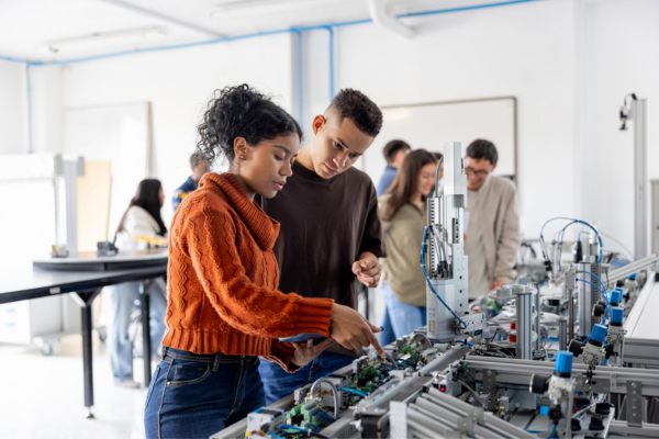 Two engineering students lean over complex robotics machinery in a bright laboratory, collaborating on an automated system while other classmates work in the background.