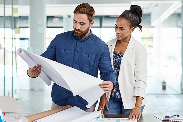 Two colleagues standing by a conference table in a bright, modern office, focused on reviewing a large architectural blueprint together. The man holds up the open roll while the woman looks on closely.