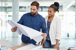 Two colleagues standing by a conference table in a bright, modern office, focused on reviewing a large architectural blueprint together. The man holds up the open roll while the woman looks on closely.