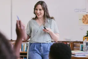 Teacher standing at the front of a classroom with a student raising their hand to ask a question
