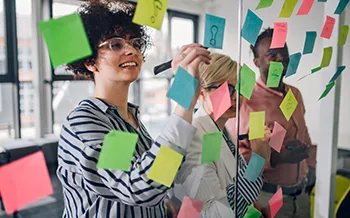 Trois personnes collaborent et discutent de notes sur un mur de verre couvert de notes autocollantes colorées dans un bureau.