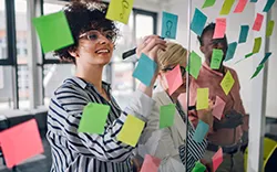 Trois personnes collaborent et discutent de notes sur un mur de verre couvert de notes autocollantes colorées dans un bureau.