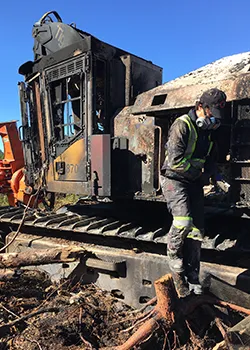 Un ouvrier en tenue réfléchissante se tient sur les voies ferrées à côté d'un wagon calciné, avec des débris et des branches d'arbres éparpillés autour.