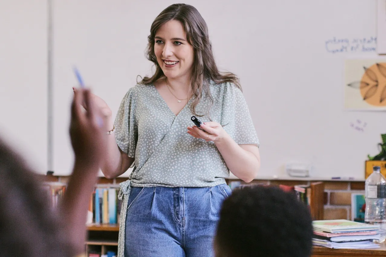 Teacher standing at the front of a classroom with a student raising their hand to ask a question
