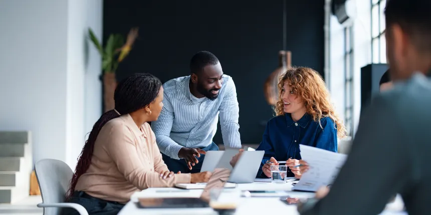 Un groupe de collègues d'origines diverses collabore dans un espace de bureau lumineux et moderne. Un homme se tient debout, penché sur une table de conférence pour discuter avec deux femmes assises, tandis que des ordinateurs portables ouverts et des documents imprimés sont étalés devant eux.