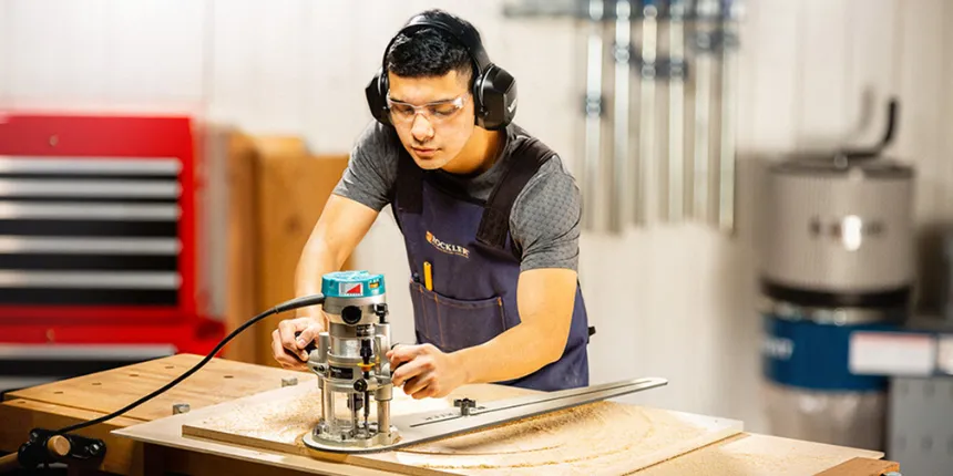 Un travailleur du bois portant des lunettes de sécurité, des protecteurs d'oreilles et un tablier utilise une défonceuse munie d'un gabarit de découpe circulaire sur un établi. De la sciure de bois recouvre la pièce de bois alors qu'il guide l'outil avec précision dans un atelier.