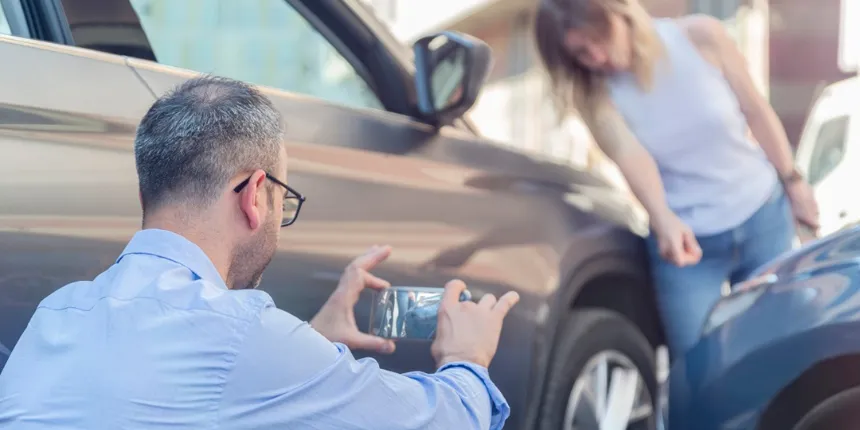 A man crouches down to take photos of vehicle damage with his smartphone after a minor car accident. A woman stands in the background looking distressed near the point of impact.