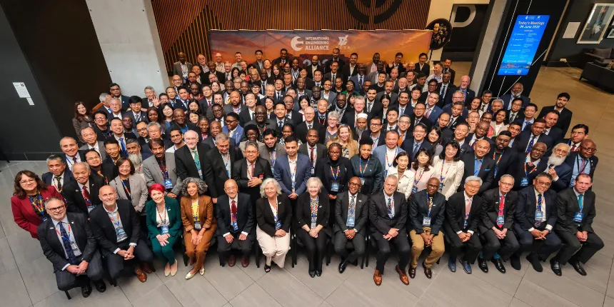 Photo de groupe réunissant des délégués rassemblés à l'intérieur pour l'événement de l'International Engineering Alliance. Les participants sont disposés en plusieurs rangées dans un atria moderne, le premier rang étant assis. Une grande bannière avec le logo de l'alliance est visible à l'arrière-plan.