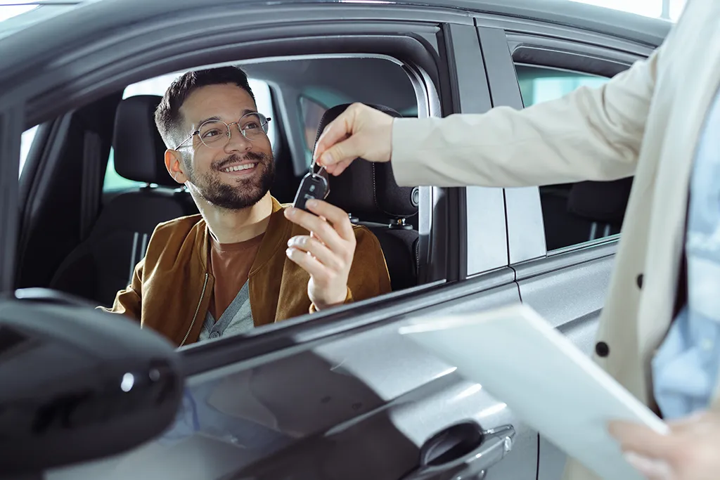 Photograph of a smiling man in eyeglasses seated behind the steering wheel of a car, receiving the car key fob from an agent holding rental paperwork outside the open driver-side window.