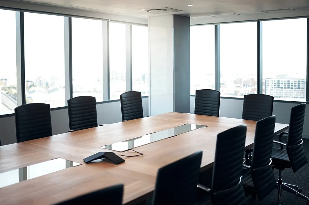 Photograph of an empty, bright modern boardroom with large floor-to-ceiling windows overlooking a city skyline. A long, light wood conference table is surrounded by high-backed black leather office chairs, with a conference phone unit positioned in the center.